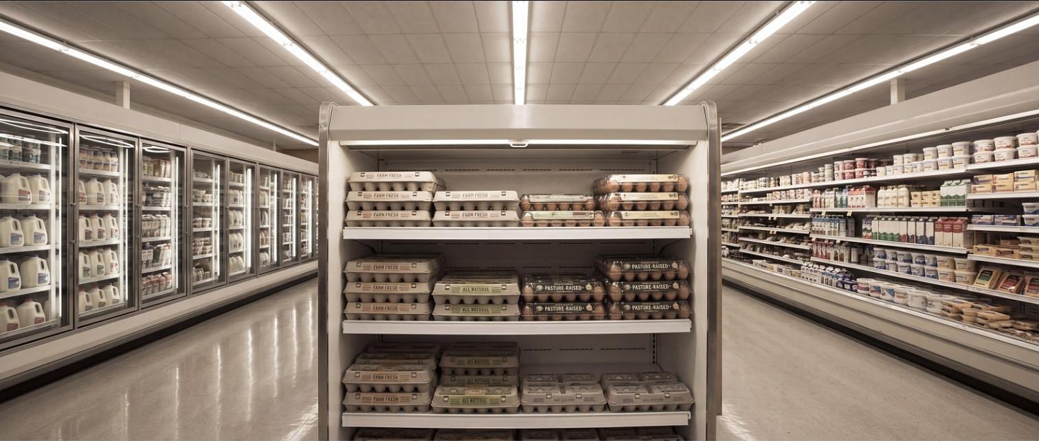 A supermarket refrigerated aisle seen head-on: a chilled case stacked with egg cartons labeled Farm Fresh, All Natural and Pasture-Raised, dairy coolers on both sides. The opening scene of the module.